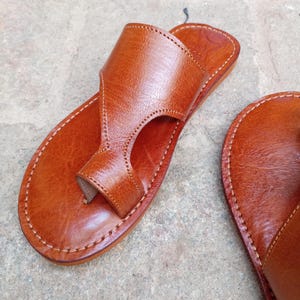 May include: Close-up of a pair of brown leather sandals. The sandals feature a thong-style design with a strap that goes over the top of the foot. The edges are stitched with white thread. The sandals are on a gray surface.