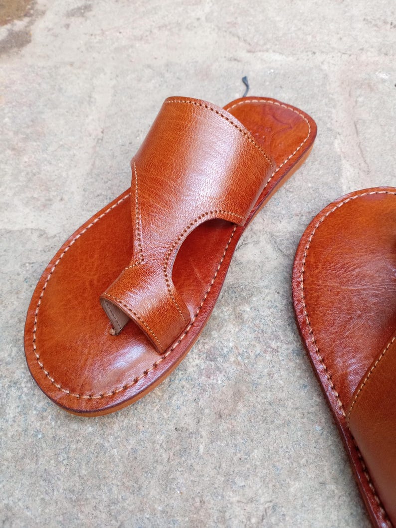 May include: Close-up of a pair of brown leather sandals. The sandals feature a thong-style design with a strap that goes over the top of the foot. The edges are stitched with white thread. The sandals are on a gray surface.