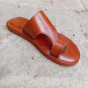 May include: A single, handcrafted leather sandal in a warm brown hue. The sandal features a toe loop and a wide strap across the top of the foot. The edges are neatly stitched, and the sole appears flat. This footwear is designed for casual wear.