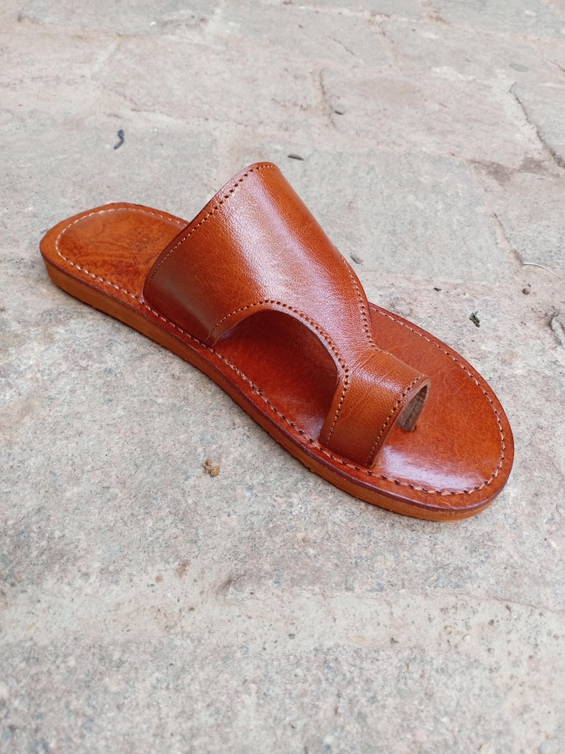 May include: A single, handcrafted leather sandal in a warm brown hue. The sandal features a toe loop and a wide strap across the top of the foot. The edges are neatly stitched, and the sole appears flat. This footwear is designed for casual wear.