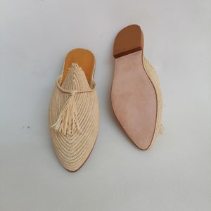 Puede incluir: Un par de zapatillas de paja beige con un detalle de borla. Las zapatillas tienen una punta puntiaguda y una suela de cuero.