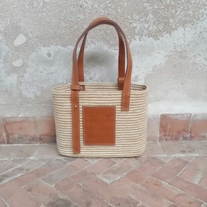 Op de afbeelding: Een beige geweven tote bag met bruine leren handvatten en een bruine leren patch aan de voorkant.