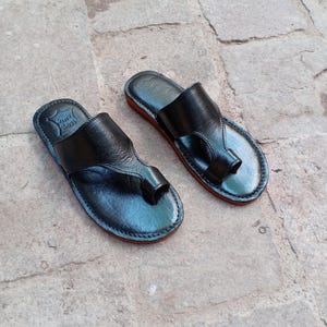 May include: A pair of black leather sandals with a toe loop design. The sandals have a red sole and are displayed on a stone surface. The sandals appear to be handmade.