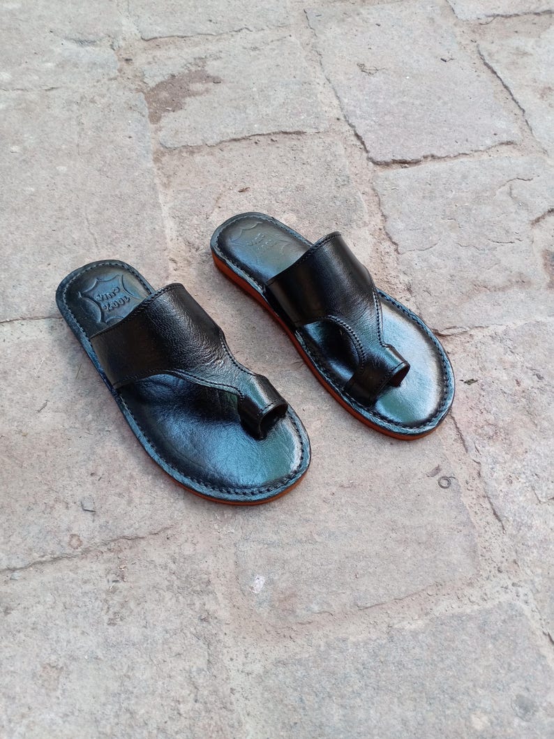 May include: A pair of black leather sandals with a toe loop design. The sandals have a red sole and are displayed on a stone surface. The sandals appear to be handmade.