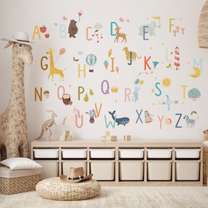 May include: A whimsical children's room scene featuring alphabet wall decals with colorful illustrations of animals and objects. A woven giraffe sculpture and storage bins add to the playful decor. The letters are in various colors, with corresponding images.