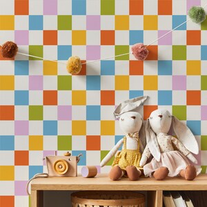 Op de afbeelding: Een witte muur met een kleurrijk geruit patroon in tinten oranje, geel, groen, blauw en roze. Twee witte knuffels van konijnen zitten op een houten plank voor de muur.