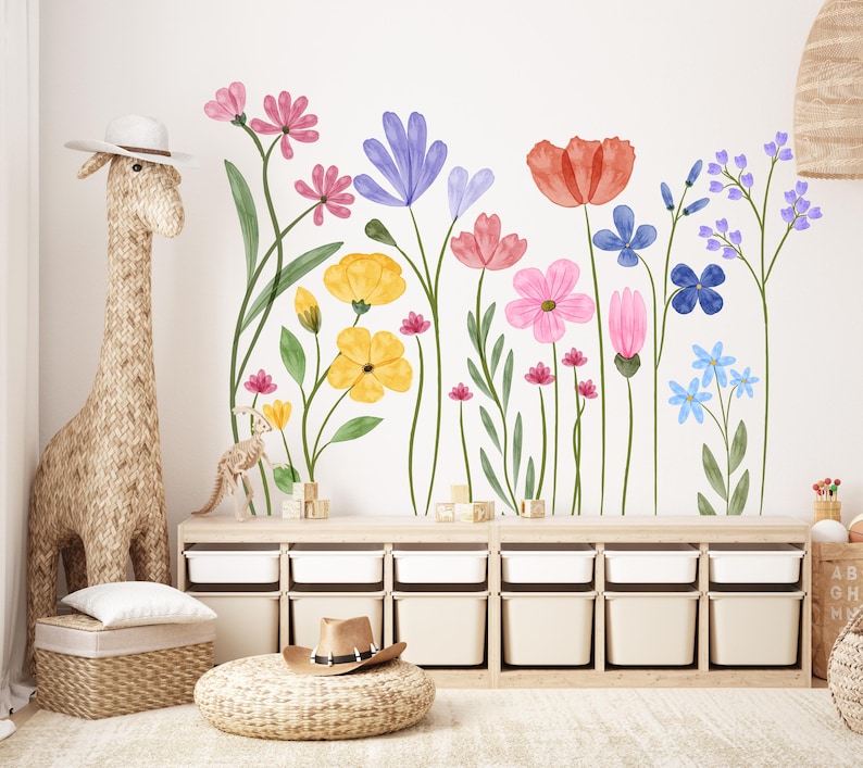 May include: A nursery scene with a giraffe-shaped storage basket, a woven ottoman with a hat, and a wall decal of colourful watercolour flowers in pink, yellow, blue, and red. A wooden storage unit with white bins is also present.