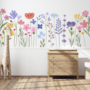 May include: A wall decal featuring a vibrant watercolour floral design with various colourful flowers, including red poppies, pink, purple, and blue blooms. A woven giraffe and a wooden dresser are in the foreground.
