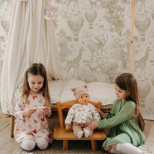 Puede incluir: Dos ni&ntilde;as jugando con una mu&ntilde;eca sentada en una peque&ntilde;a silla de madera. La mu&ntilde;eca lleva un conjunto beige y un sombrero. Las ni&ntilde;as est&aacute;n en una habitaci&oacute;n con papel pintado con tem&aacute;tica de safari y una peque&ntilde;a tienda de campa&ntilde;a.
