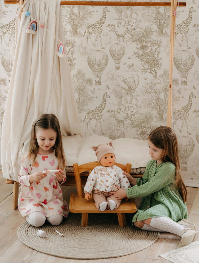 Puede incluir: Dos ni&ntilde;as jugando con una mu&ntilde;eca sentada en una peque&ntilde;a silla de madera. La mu&ntilde;eca lleva un conjunto beige y un sombrero. Las ni&ntilde;as est&aacute;n en una habitaci&oacute;n con papel pintado con tem&aacute;tica de safari y una peque&ntilde;a tienda de campa&ntilde;a.
