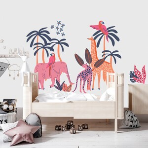 Op de afbeelding: Een muursticker-set met een roze olifant met een papegaai, een giraf en een luipaard tussen donkerblauwe palmbomen en tropische planten. Het ontwerp bevat kleine blauwe bloemen en is afgebeeld op een witte muur boven een lichthouten bedframe.