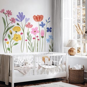 May include: A white cot with a neutral blanket and pillows is in a room with a floral wall mural. The mural features colourful watercolour flowers in shades of pink, yellow, blue, and red. A woven basket and a rug complete the nursery setting.