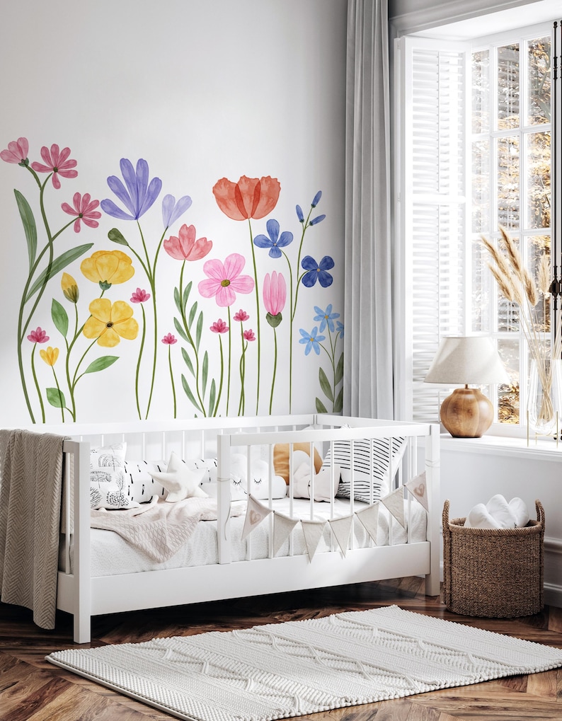 May include: A white cot with a neutral blanket and pillows is in a room with a floral wall mural. The mural features colourful watercolour flowers in shades of pink, yellow, blue, and red. A woven basket and a rug complete the nursery setting.