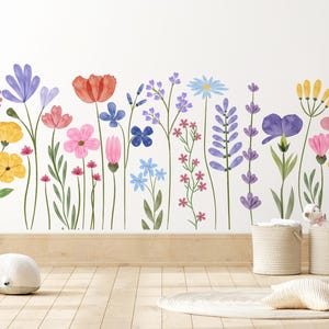 May include: A vibrant floral wall sticker showcasing watercolour flowers in pink, purple, yellow, and blue. The sticker is on a white wall, with a wooden floor and decorative items in the foreground. The flowers are in full bloom.