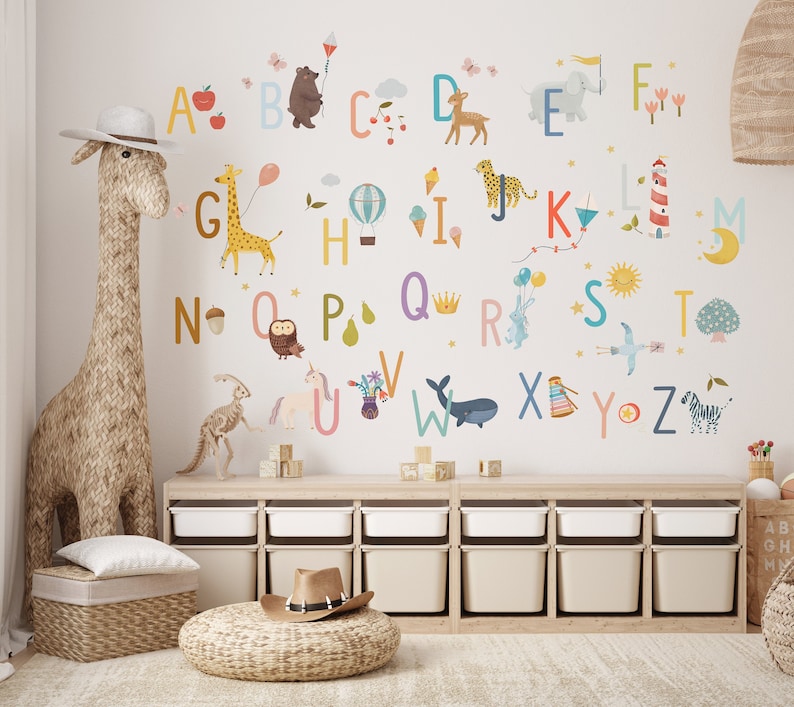 May include: A whimsical children's room scene featuring alphabet wall decals with colorful illustrations of animals and objects. A woven giraffe sculpture and storage bins add to the playful decor. The letters are in various colors, with corresponding images.