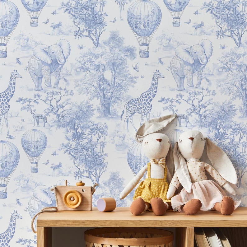 May include: A wooden toy camera and a wooden cylinder sit on a light wood shelf in front of a blue and white toile wallpaper. Two stuffed animal dolls are also on the shelf. The wallpaper features elephants, giraffes, hot air balloons, and trees.