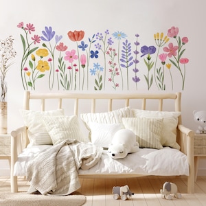 May include: A wall decal of watercolour flowers in pink, yellow, blue, and purple shades. The flowers are above a light wooden bench with white cushions and a blanket. A stuffed polar bear and elephant are on the bench. The scene is set indoors.