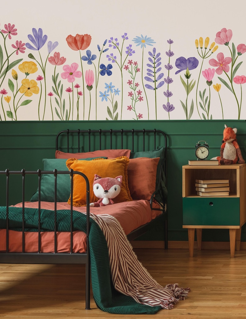 May include: A bedroom with a black metal bed, rust-coloured bedding, orange and green cushions, and a green blanket. A fox toy sits on the bed. The wall features a floral mural with watercolour flowers. A wooden bedside table with a green drawer and a clock is visible.