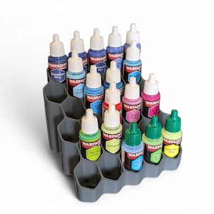 May include: A gray plastic paint bottle organizer filled with various colors of paint bottles. The bottles are arranged in a stepped configuration, showcasing a range of colors including blue, pink, green, and yellow. The bottles have white caps and labels.