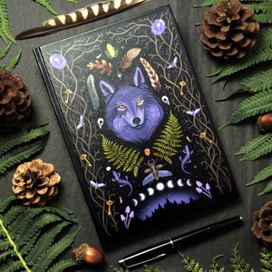 Puede incluir: Un diario negro con una ilustración de un lobo morado rodeado de imágenes de bosque, incluyendo helechos, ramas y fases lunares. La portada presenta un diseño detallado con detalles dorados.