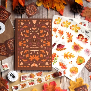 May include: A brown journal with a colourful autumn leaf design and the text "You are amazing!" on the cover. The journal is surrounded by autumn-themed stickers, washi tape, and decorative leaves.