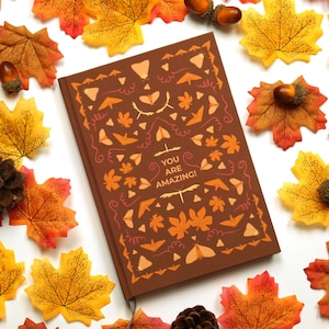 Könnte beinhalten: Ein braunes Notizbuch mit einem Herbst-Design mit orangefarbenen, gelben und roten Blättern und dem Text "You are amazing!" auf dem Einband.