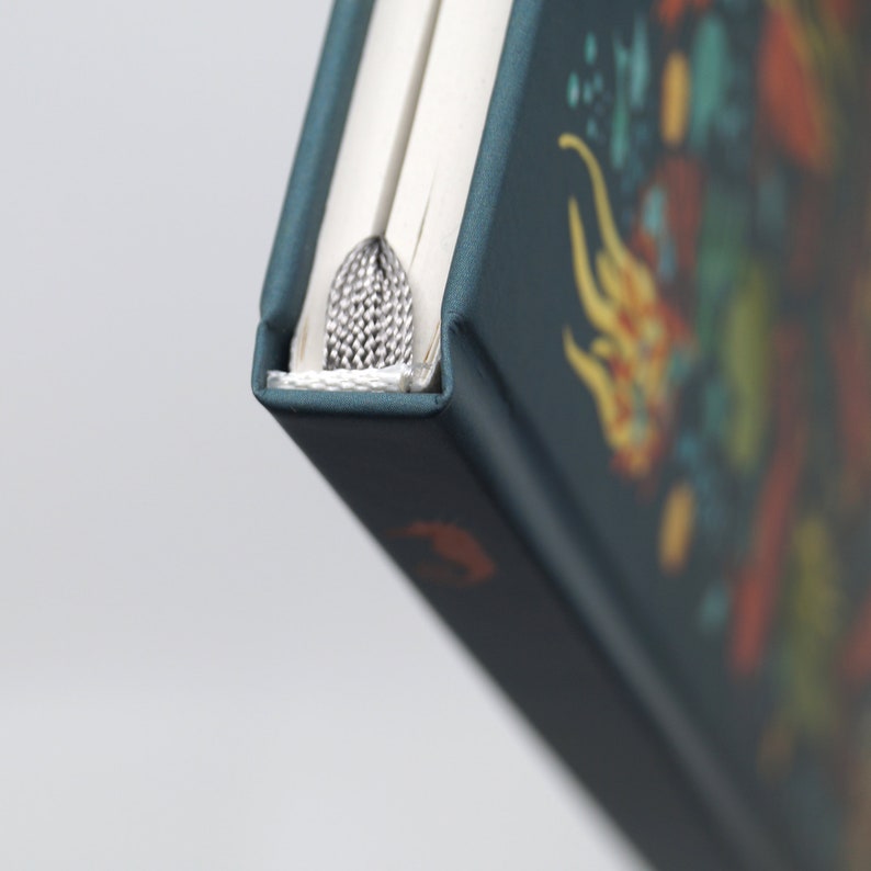 May include: A teal-colored journal with a decorative design featuring yellow, orange, and blue elements. The journal has a silver bookmark and white-paged interior. The cover is a dark teal color.