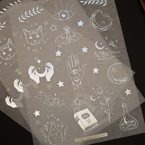 K&ouml;nnte beinhalten: Ein Blatt transparentes Stickerpapier mit wei&szlig;en Illustrationen von Monden, Sternen, H&auml;nden, Katzen, einem Nachtfalter, einer Schlange, einem Buch und Flaschen. Die Sticker sind perfekt, um Tageb&uuml;chern, Planern und anderen Papierarbeiten einen Hauch von Verspieltheit zu verleihen.