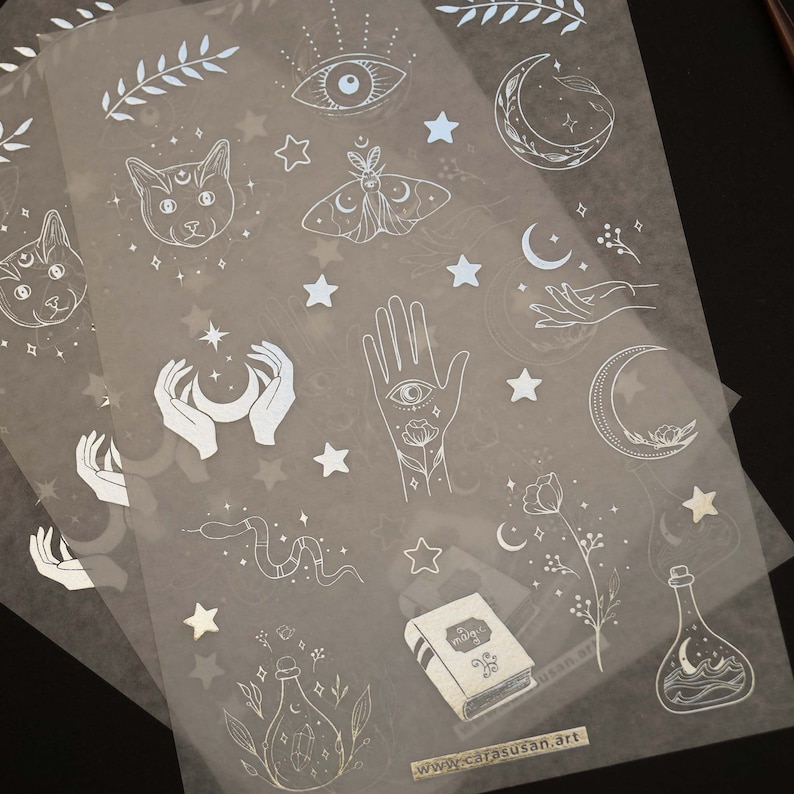K&ouml;nnte beinhalten: Ein Blatt transparentes Stickerpapier mit wei&szlig;en Illustrationen von Monden, Sternen, H&auml;nden, Katzen, einem Nachtfalter, einer Schlange, einem Buch und Flaschen. Die Sticker sind perfekt, um Tageb&uuml;chern, Planern und anderen Papierarbeiten einen Hauch von Verspieltheit zu verleihen.