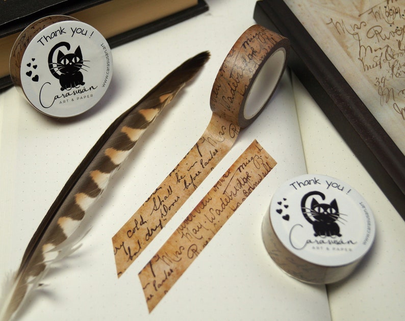 May include: Two rolls of brown washi tape with black and white illustrations of a cat and handwritten text. The text on the tape reads "Cold Shall be in" and "7 Was hay. Wadebridge. B. Cornwall". The rolls of tape are on a white surface with a brown feather.