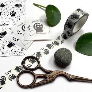 Puede incluir: Un rollo de cinta washi blanco y negro con un patr&oacute;n repetitivo de gatos negros. La cinta est&aacute; parcialmente desenrollada y hay unas tijeras, una peque&ntilde;a piedra y una pegatina con el texto "&iexcl;Gracias! Cara Susan Art & Paper" sobre un fondo blanco.