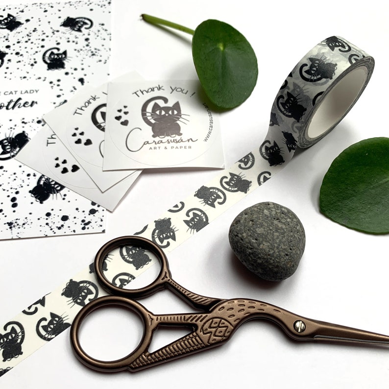 Puede incluir: Un rollo de cinta washi blanco y negro con un patr&oacute;n repetitivo de gatos negros. La cinta est&aacute; parcialmente desenrollada y hay unas tijeras, una peque&ntilde;a piedra y una pegatina con el texto "&iexcl;Gracias! Cara Susan Art & Paper" sobre un fondo blanco.