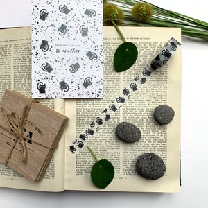 Puede incluir: Una tarjeta blanca con siluetas de gatos negros y el texto "FROM ONE CAT LADY to another". La tarjeta est&aacute; sobre un libro con texto en blanco y negro, hojas verdes y cinta washi en blanco y negro. Tambi&eacute;n hay tres rocas volc&aacute;nicas negras.