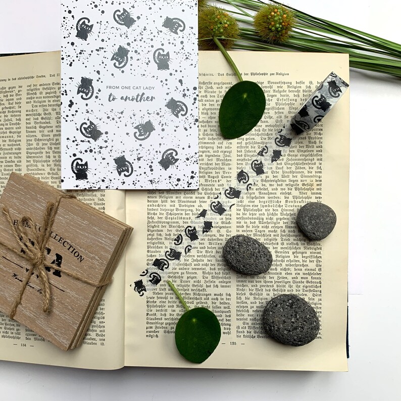 Puede incluir: Una tarjeta blanca con siluetas de gatos negros y el texto "FROM ONE CAT LADY to another". La tarjeta est&aacute; sobre un libro con texto en blanco y negro, hojas verdes y cinta washi en blanco y negro. Tambi&eacute;n hay tres rocas volc&aacute;nicas negras.