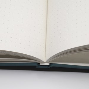 May include: Open notebook with dot grid pages. The pages are off-white and have small gray dots arranged in a grid pattern. The notebook has a teal cover and a white ribbon bookmark. The notebook is open, revealing the interior pages.