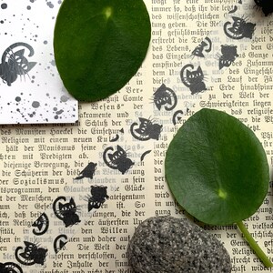 Puede incluir: Silueta negra y blanca de un gato repetida sobre un fondo blanco con puntos negros. El fondo es una p&aacute;gina de un libro con texto alem&aacute;n. Hay dos hojas verdes y una piedra gris en la p&aacute;gina.