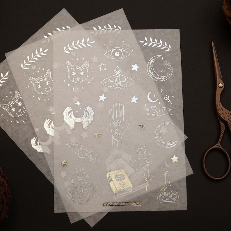 K&ouml;nnte beinhalten: Drei Bl&auml;tter transparentes Aufkleberpapier mit silbernen Folienmotiven. Die Motive umfassen eine Katze, einen Nachtfalter, einen Halbmond, eine Hand mit einem Auge, ein Buch, eine Flasche und Sterne.