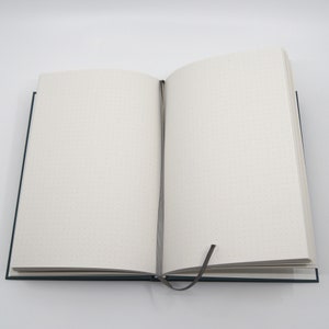 May include: An open, teal-colored journal with dot grid pages. A gray ribbon bookmark is inserted between the pages. The journal is on a white background.