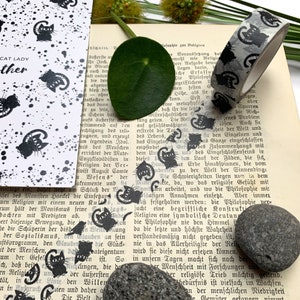 Puede incluir: Una hoja de papel con estampado en blanco y negro con un dise&ntilde;o de gato negro. El papel est&aacute; al lado de un libro con cinta washi estampada en blanco y negro. El libro tiene texto en alem&aacute;n. Hay una peque&ntilde;a hoja verde y una piedra gris en la imagen.