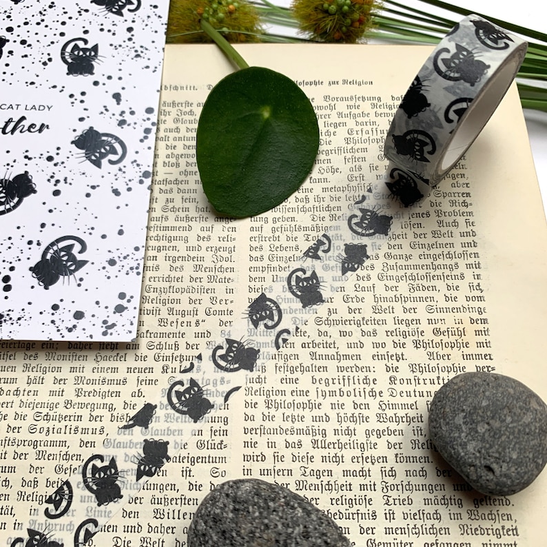 Puede incluir: Una hoja de papel con estampado en blanco y negro con un dise&ntilde;o de gato negro. El papel est&aacute; al lado de un libro con cinta washi estampada en blanco y negro. El libro tiene texto en alem&aacute;n. Hay una peque&ntilde;a hoja verde y una piedra gris en la imagen.