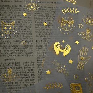 K&ouml;nnte beinhalten: Goldfolien-Aufkleber mit mystischem Thema. Die Aufkleber zeigen verschiedene Designs, darunter eine Katze, einen Halbmond, Sterne, ein Auge, einen Schmetterling und H&auml;nde, die einen Halbmond halten.