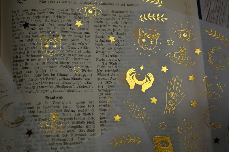 K&ouml;nnte beinhalten: Goldfolien-Aufkleber mit mystischem Thema. Die Aufkleber zeigen verschiedene Designs, darunter eine Katze, einen Halbmond, Sterne, ein Auge, einen Schmetterling und H&auml;nde, die einen Halbmond halten.