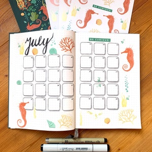 May include: Open bullet journal spread for July with seahorse and coral illustrations. The word "July" is handwritten in black ink. The spread includes a calendar grid with the text "BE CURIOUS!" and decorative stickers.