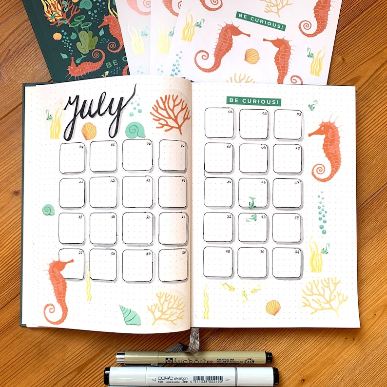 May include: Open bullet journal spread for July with seahorse and coral illustrations. The word "July" is handwritten in black ink. The spread includes a calendar grid with the text "BE CURIOUS!" and decorative stickers.