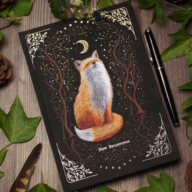 Witchy notebook "New Beginnings" with hand-painted fox illustration, gold foil moon phases and stars on black hardcover, A5 journal with Munken Pure paper. Cover view.