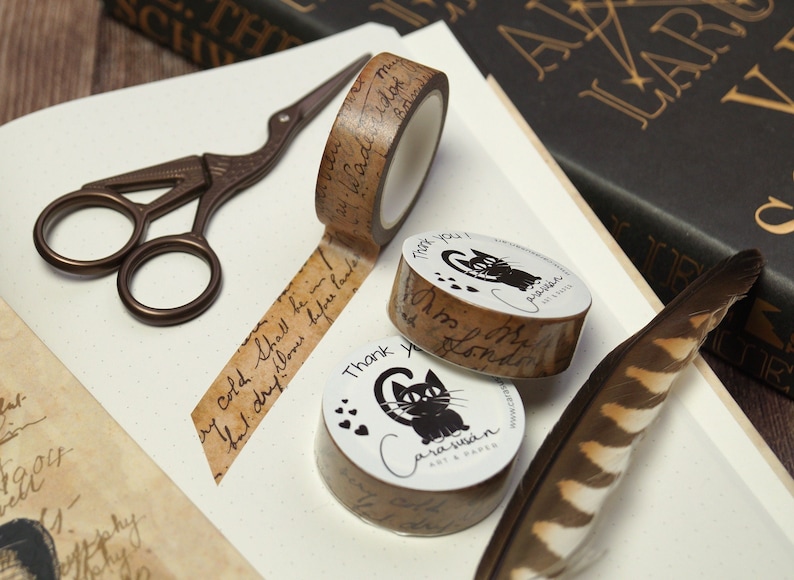May include: Three rolls of decorative washi tape with a vintage aesthetic. The tape features a brown background with black handwritten text and illustrations of cats. The text on the tape reads "Thank you! I'm so London" and "Carasusan Art & Paper".