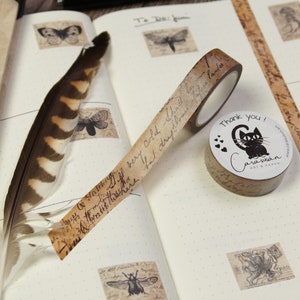 May include: A roll of brown washi tape with a black cat design and the text "Thank you!"  The tape is on a white notebook with other decorative elements.