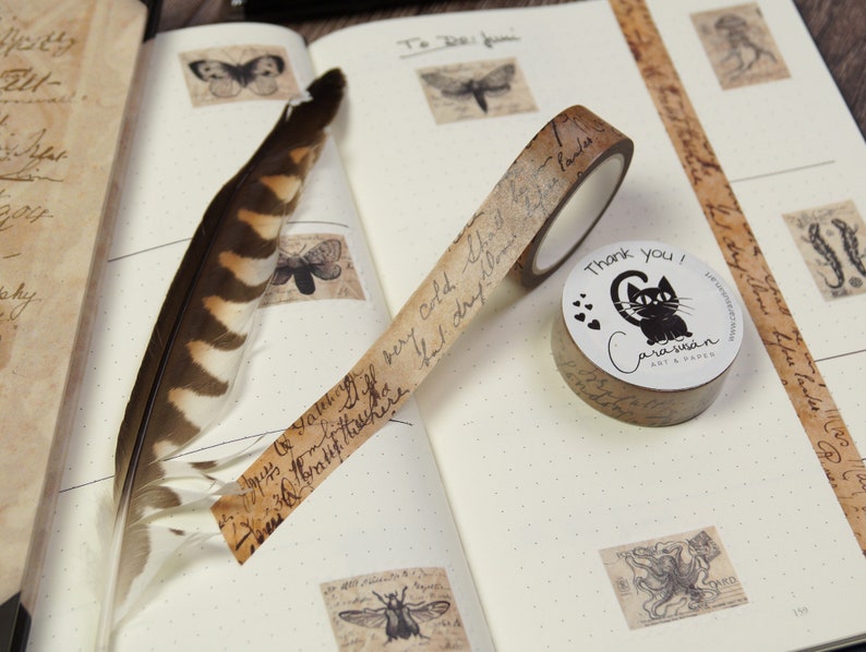 May include: A roll of brown washi tape with a black cat design and the text "Thank you!"  The tape is on a white notebook with other decorative elements.