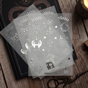 K&ouml;nnte beinhalten: Drei Bl&auml;tter transparentes Stickerpapier mit silbernen Folienmotiven. Die Motive umfassen Katzen, Monde, Sterne, H&auml;nde, Augen und einen Schmetterling. Die Sticker sind perfekt, um Tageb&uuml;chern, Planern und anderen Bastelprojekten einen Hauch von Magie zu verleihen.