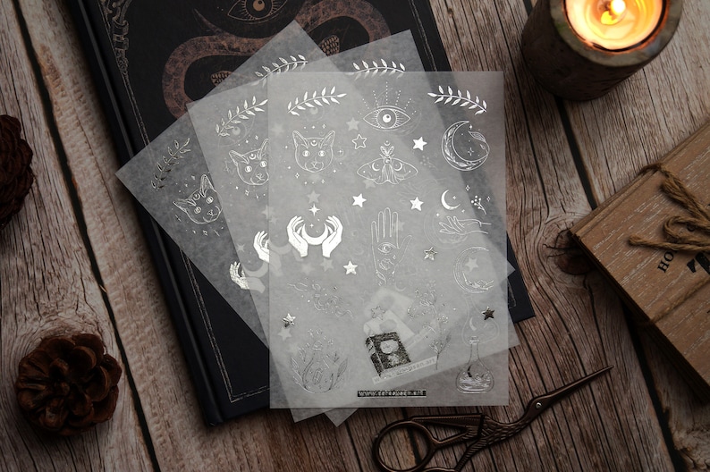 K&ouml;nnte beinhalten: Drei Bl&auml;tter transparentes Stickerpapier mit silbernen Folienmotiven. Die Motive umfassen Katzen, Monde, Sterne, H&auml;nde, Augen und einen Schmetterling. Die Sticker sind perfekt, um Tageb&uuml;chern, Planern und anderen Bastelprojekten einen Hauch von Magie zu verleihen.